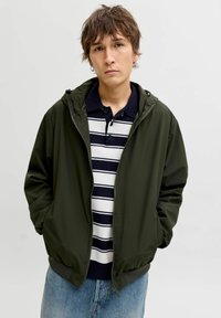 Olivengrøn lynlåstrøje med hætte, glat stof, ribstrikkede manchetter, lagdelt over en navyblå og hvid stribet polo, kombineret med lyse denimjeans.