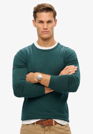 Dunkelgrüner, gestrickter Rundhals-Pullover mit gerippter Textur, weißem Innencollar und Logoakzent am linken Ärmel. Camelfarbener Gürtel und Uhr sind ebenfalls enthalten.