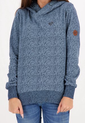 Nainen, joka pukeutunut siniseen kuviolliseen huppariin, jossa on pieniä sydänkuvioita ja resorihihat, yhdistettynä sinisiin farkkuihin, seisoo valkoista taustaa vasten.