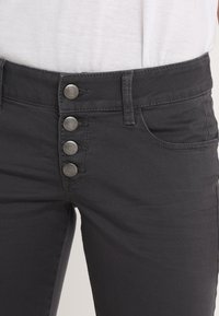 Jean en denim noir à coupe ajustée, avec une taille haute et quatre boutons métalliques au niveau de la fermeture. Inclut une poche avant.