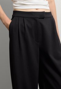 Pantalones de tela negra con pliegues que presentan un tejido suave, cintura alta, bolsillos laterales y una silueta limpia y ajustada.