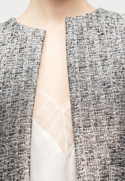 Struktuurne must-valge jakk, millel on madal kaelus, paljastab selle all pehme, heledat tooni kampsuni. Jakil on puhas, avatud esiosa.