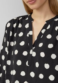 Zwarte shirt met V-hals, lange mouwen en een patroon van witte cirkelvormige stippen. De stof lijkt lichtgewicht met een gestructureerde afwerking.