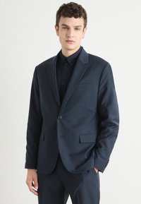 Abito blu navy con rever a punta e chiusura a un bottone. Presenta tasche dritte e una texture liscia, abbinato a una camicia scura.