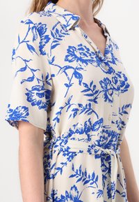 Šaty s krátkým rukávem a límečkem s modrým květinovým vzorem na krémovém podkladu, knoflíková přední část a zavazovací pás.
