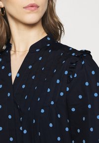 Marinblus med blåa prickar, med rynkad axeldetalj och veck längs fram. Tyget ser slätt och lätt ut.