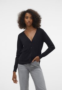 Cardigan negru cu decolteu în V, închidere cu nasturi și mâneci lungi, purtat peste blugi gri. Textură netedă, design minimalist.
