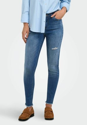 Personne portant une chemise bleu clair boutonnée, un jean bleu ajusté avec un léger effet usé, des ourlets effilochés et des mocassins en daim marron, debout avec une main dans la poche.