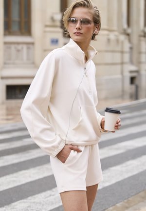 Felpa bianca con zip, dal taglio morbido e con tasche, abbinata a pantaloni corti coordinati. Il modello tiene in mano una tazza di caffè usa e getta. Ambiente urbano.