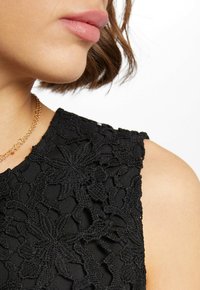 Zwarte mouwloze top met verfijnde kantdetails en bloemmotieven. Glad oppervlak en een ronde halslijn, geaccentueerd met een gouden ketting.