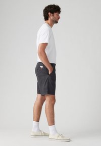 Pantaloni scurți gri dintr-un amestec de bumbac, cu buzunare laterale și un patch cu logo pe spate, purtați cu un tricou alb și adidași cu tălpi joase.