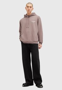 Lys mauve hættebluse med snørebånd og frontlomme, parret med sort vidde jeans og sorte platform sko. Simpelt logo detalje på brystet.