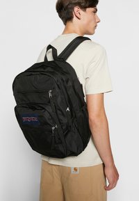 La mochila negra Jansport cuenta con múltiples compartimentos con cremallera, un bolsillo lateral de malla y correas de hombro acolchadas. El material es de tela resistente.