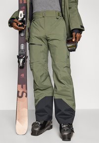 Olive žaliai spalvos slidinėjimo kelnės su juodais akcentais, turinčios užtrauktukus ir kišenes. Slidinėjimo įranga apima juodus batus ir slides su tvirtinimais.