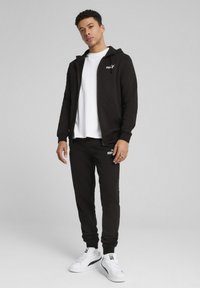 Felpa nera con zip frontale e due tasche, abbinata a pantaloni jogger neri coordinati. Accenti di logo bianchi su entrambi, abbinati a sneakers bianche.