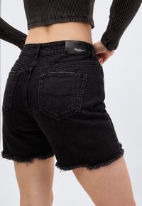 Γυναίκα που φοράει μαύρο denim σορτς με φθαρμένα τελειώματα και εφαρμοστό μακρυμάνικο ριμπ crop top, με τα χέρια της ακουμπισμένα στους γοφούς.