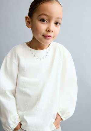 Mlada djevojka s glatkom kosom, nosi bijelu teksturiranu bluzu i nježnu ogrlicu od perlica, stoji na sivom obliku pozadine.