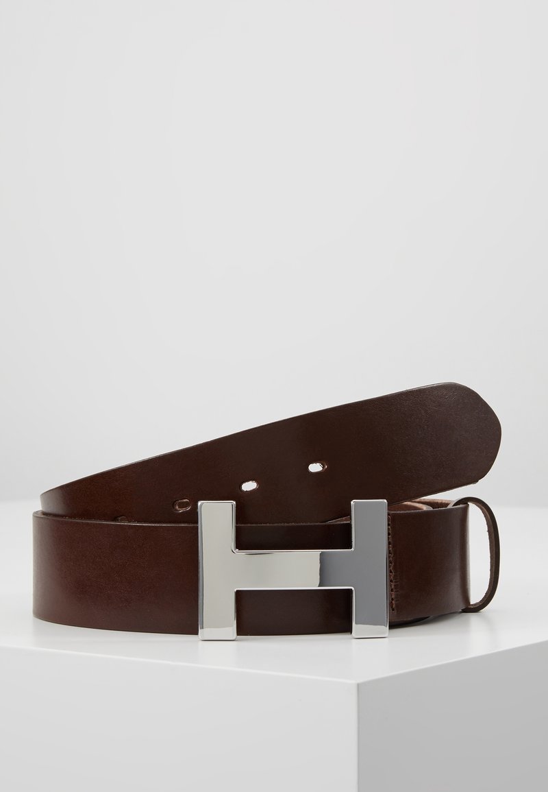 Ceinture en cuir marron enroulée sur une surface blanche avec une grande boucle argentée en forme de H tournée vers l'avant.