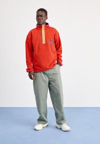 Mand står med hænderne i lommerne iført en orange fleecepullover, lysegrå bukser og hvide sneakers på en blå gulv mod en hvid væg.