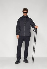 Casaco impermeável preto com fecho de correr à frente, gola alta e bolsos com fecho, combinado com calças a condizer e bastões de trekking. Calçado robusto completa o visual.