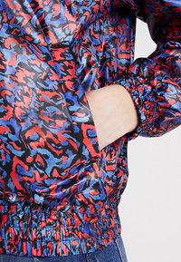 Veste doudoune colorée avec une base noire et un motif abstrait rouge et bleu, manches élastiques et taille froncée.
