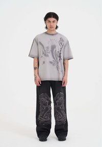 Camiseta gris de gran tamaño con gráficos de águila y logo, combinada con pantalones vaqueros negros con bordados de dragón y detalles desgastados.