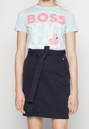 Kvinde iført en lyseblå T-shirt med teksten "BOSS BEACH" og pink flamingo-grafik, kombineret med et sort, højtaljet skørt med bundet bælte.