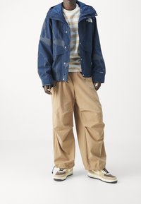 Modrá voděodolná bunda s kapucí a šedými akcenty, v kombinaci s béžovými volnými kalhotami a hnědými teniskami s bílými a žlutými detaily.