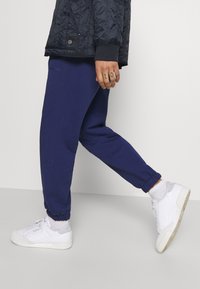 Marinblå mjuka byxor med elastiska muddar, kompletterade med en svart vadderad jacka. Vita sneakers med grå detaljer. Mjuk bomullsblandad textur.