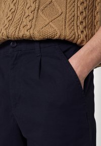 Chinos blu navy realizzati in cotone con una texture liscia, dotati di chiusura a bottone e tasche laterali, abbinati a un maglione marrone zigrinato.