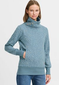 Sudadera de color teal con cuello alto, que cuenta con tres botones, diseñada con patrones de puntos en tonos claros y oscuros dispersos, y bolsillos delanteros.