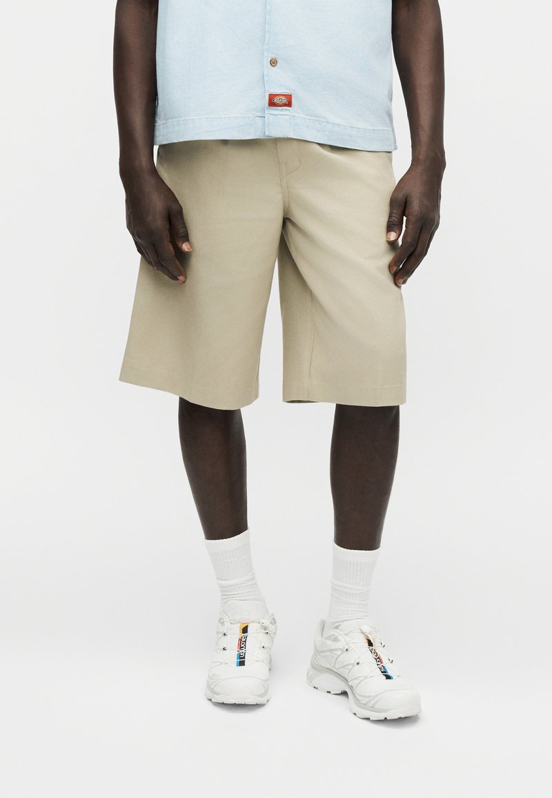 Homme portant un short beige mi-cuisses, une chemise bleu clair, des chaussettes blanches et des baskets blanches avec des bandes colorées sur un fond uni.