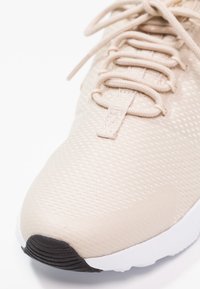 Beige idrottssko med nätöverdel, platta snören, texturerad yta, rund tå och svart gummisula.