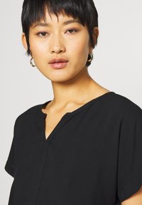 Musthave muströöpne top musta värviga, millel on v-kujuline kaelus ja lõdvestunud istuvus. Valmistatud pehmest kangast, millel on sile tekstuur ja minimaalne kaunistus.