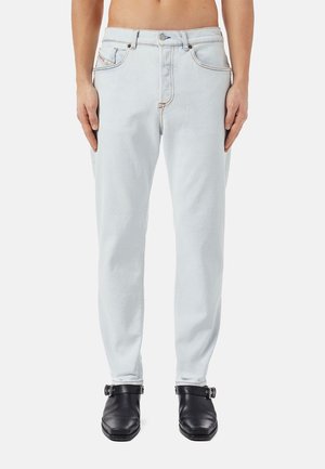 Mand iført lyseblå jeans og sorte læderankelstøvler, stående med armene afslappet langs siden mod en hvid baggrund.