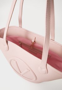 Rosa lærhåndveske med teksturert ytre, som har en stor logo på forsiden, to håndtak og en kontrastfarget rosa innside med en glidelomme.