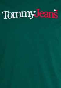 Camiseta de algodón verde con el logo de "Tommy Jeans" en blanco y rojo. Diseño simple, cuello redondo y textura suave. Sin patrones ni acentos adicionales.