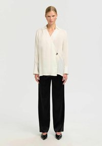 Weißes Wickelshirt mit langen Ärmeln, mit einem spitzen Kragen und einem dekorativen Knopf. Kombiniert mit hochtaillierten, schwarzen, weit geschnittenen Hosen.