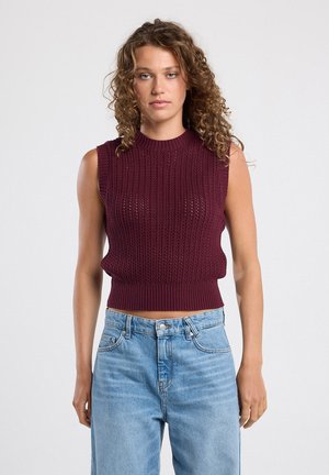 Burgunderfarbener gestrickter ärmelloser Pullover mit gerippter Textur und hohem Kragen, kurz geschnitten. Kombiniert mit hellblauen hoch taillierten Jeans.