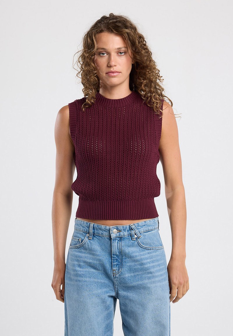 Burgundin värinen neule ilman hihoja, jossa on ribbikuvio ja korkea kaula-aukko, lyhyt malli. Yhdistetty vaaleansinisiin korkea vyötäröön sopiviin farkkuihin.