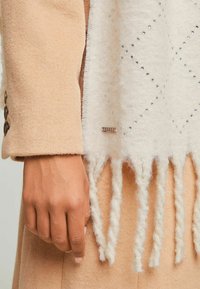 Crème sjaal met een ruitpatroon en franjes, gecombineerd met een beige stoffen jas met grote knopen en een gladde textuur. Metalen merklabel zichtbaar.
