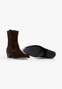 Bruine suedekampen met spitse neuzen, zichtbare stiksels en een lage blokhak. Eén laars is van de zijkant te zien, de andere van onderen.