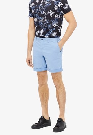 Mann trägt schwarzes, kurzärmeliges Hemd mit Blumenmuster, hellblaue Shorts mit aufgekrempeltem Saum und schwarze Sneaker, Hand in der Hosentasche, vor weißem Hintergrund.