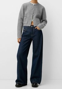 Graue, gestrickte, kurze Strickjacke mit großen Knöpfen, kombiniert mit dunkelblauen, weiten Jeans und schwarzen, klobigen Schuhen. Glatter Stoff und klare Linien.