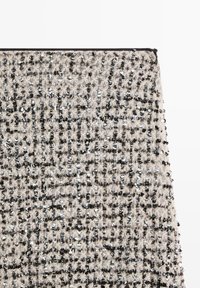 Gestructureerde stof in beige en zwart, met een roosterpatroon en metalen pailletten voor extra detail. De randen zijn afgewerkt met een zwarte bies.