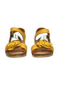 Sandalias amarillas con tiras en cruz plateadas y un acento decorativo de flor en el dedo del pie. Presentan una suela de corcho texturizada y un acabado suave.