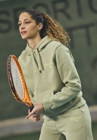 Hoodie atlético verde claro com logotipo, combinado com calças de moletom a condizer. Segurando uma raquete de ténis, tecido texturizado, ajuste relaxado.