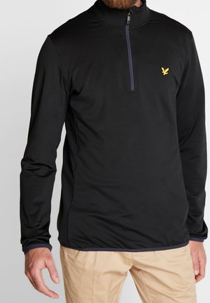 Mężczyzna ubrany w czarną bluzy z długim rękawem i suwakiem do połowy, z żółtym logo ptaka na piersi oraz beżowe spodnie, stoi na jednolitym tle.