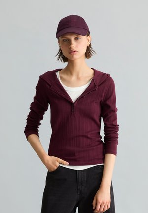 Jeune femme portant un sweat à capuche bordeaux par-dessus une chemise blanche, un pantalon noir et une casquette de baseball bordeaux assortie, posant avec une main dans la poche.