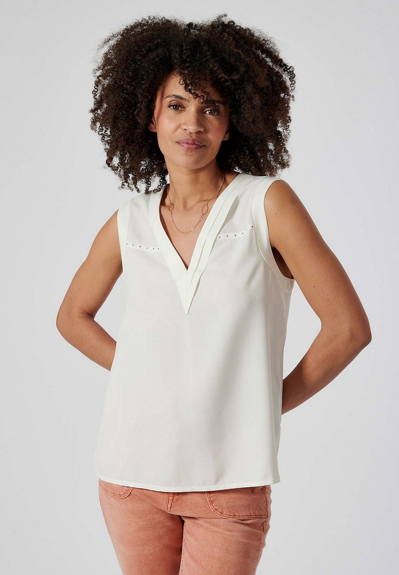 Débardeur blanc sans manches avec un col en V, présentant des découpes subtiles et une texture lisse, associé à un pantalon rose clair.